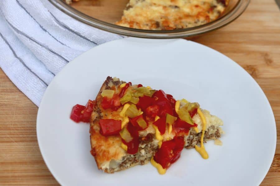 slice of cheeseburger pie topped with ketchup, mustard, pickle and tomato on a white plate