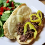 shredded beef topped with peperoncini peppers on a hoagie bun. Salad in background on a white plate.