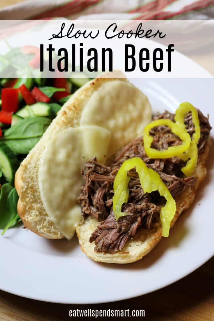 Italian beef hoagie sandwich topped with peppers on a white plate. Salad in the background.