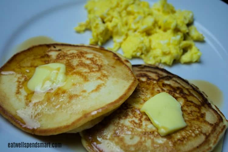 two pancakes with butter and syrup with eggs in the background on a white plate