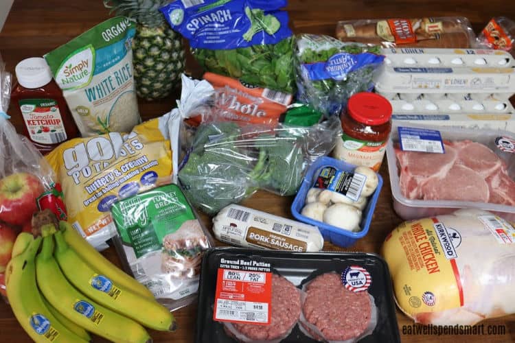 groceries on a wooden countertop: chicken, pork chops, burgers, eggs, bananas, apples, potatoes, ketchup, rice, pineapple, spinach, broccoli, mushrooms, and pasta sauce