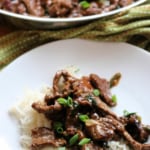 Mongolian beef on top of rice on a white plate. Skillet of Mongolian beef in the background.