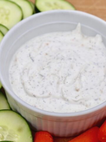 Sour cream dip in a white bowl surrounded by sliced cucumbers and baby carrots