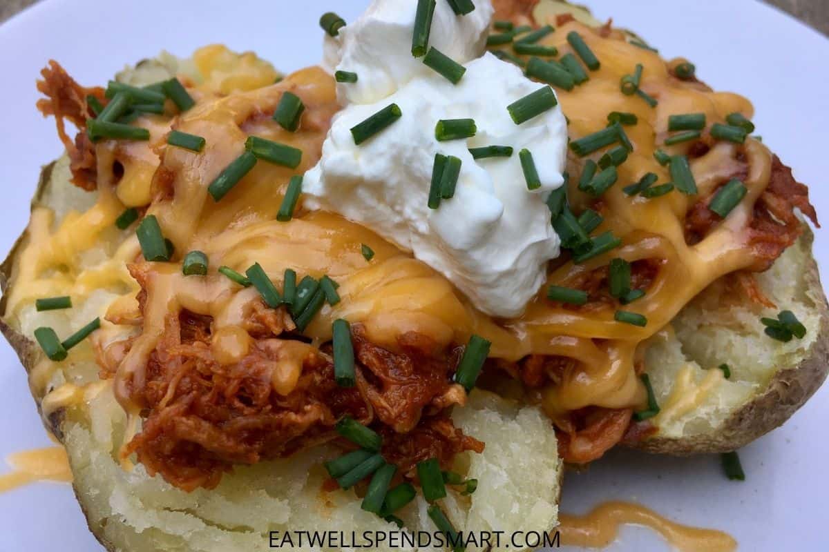 slow cooker bbq chicken thighs on a baked potato topped with cheese, sour cream and chives