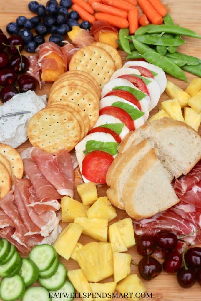 Wooden board filled with cured meats, berries, pineapple, cherries, cucumber, carrots, caprese, goat cheese, crackers, and bread
